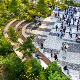 A view of a brick courtyard full of people gathering around standing tables, with trees interspersed throughout the courtyard space and a rich border of terraced cement seating and planted landscapes