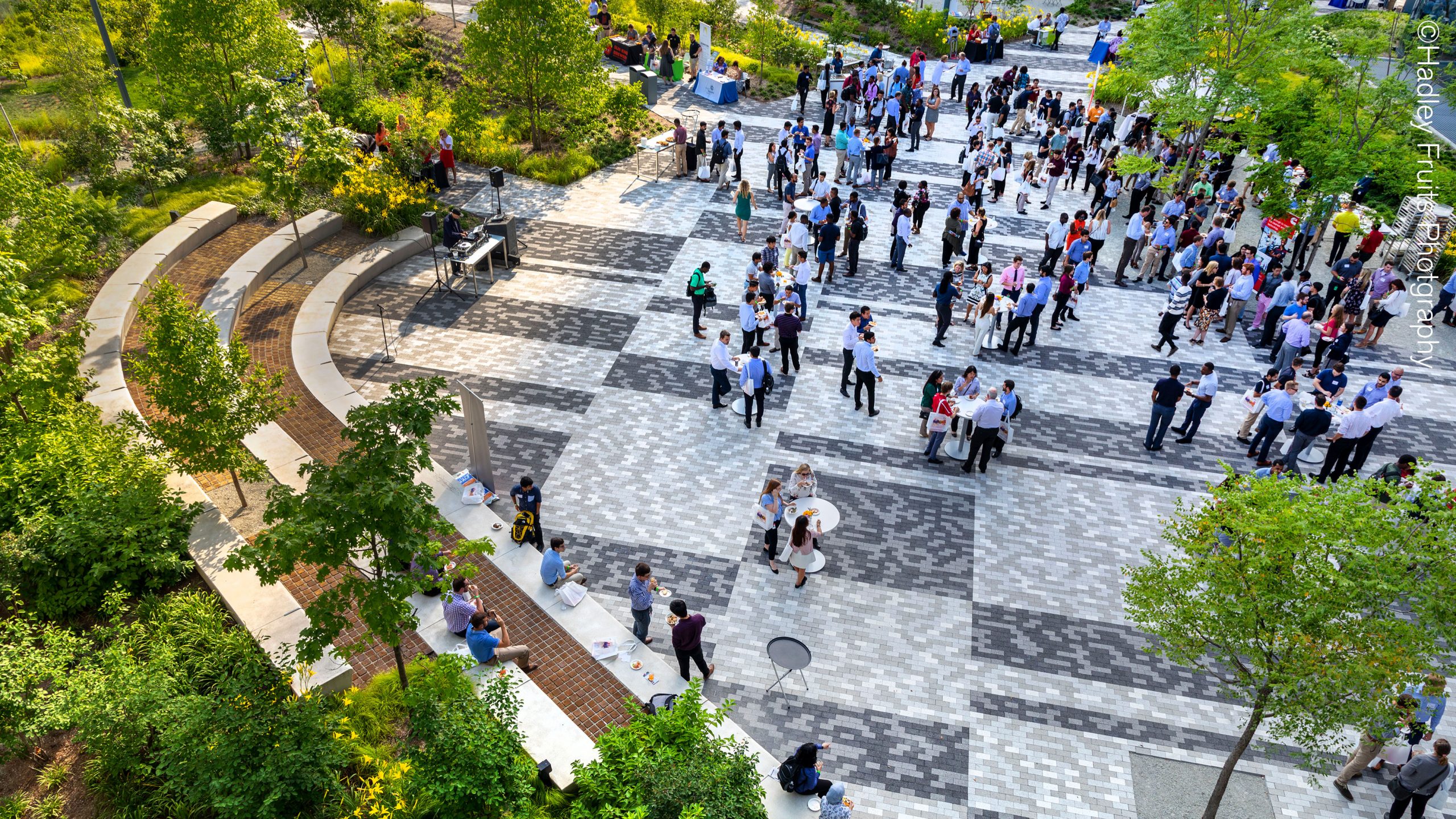 A view of a brick courtyard full of people gathering around standing tables, with trees interspersed throughout the courtyard space and a rich border of terraced cement seating and planted landscapes