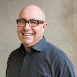 A man with a completely shaved head, rectangular glasses, and a buttoned black shirt poses for a headshot, his lips upturned