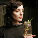 A woman with dark, wavy hair poses for a headshot with a glass jar full of water and plant clippings; she is wearing a black dress bordered with black sequins