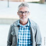 A man with short-cropped gray hair, rectangular glasses, stubble, and a blue shirt and jacket poses for a headshot