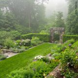 A gray stone archway bordered by tall green hedges frames a quiet private garden space, full of blooming alliums and flowering shrubs