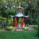 A pagoda structure in red and bronze sits atop a round dais of stone, surrounded by a water feature of lotus plants, cascades, and weeping willows