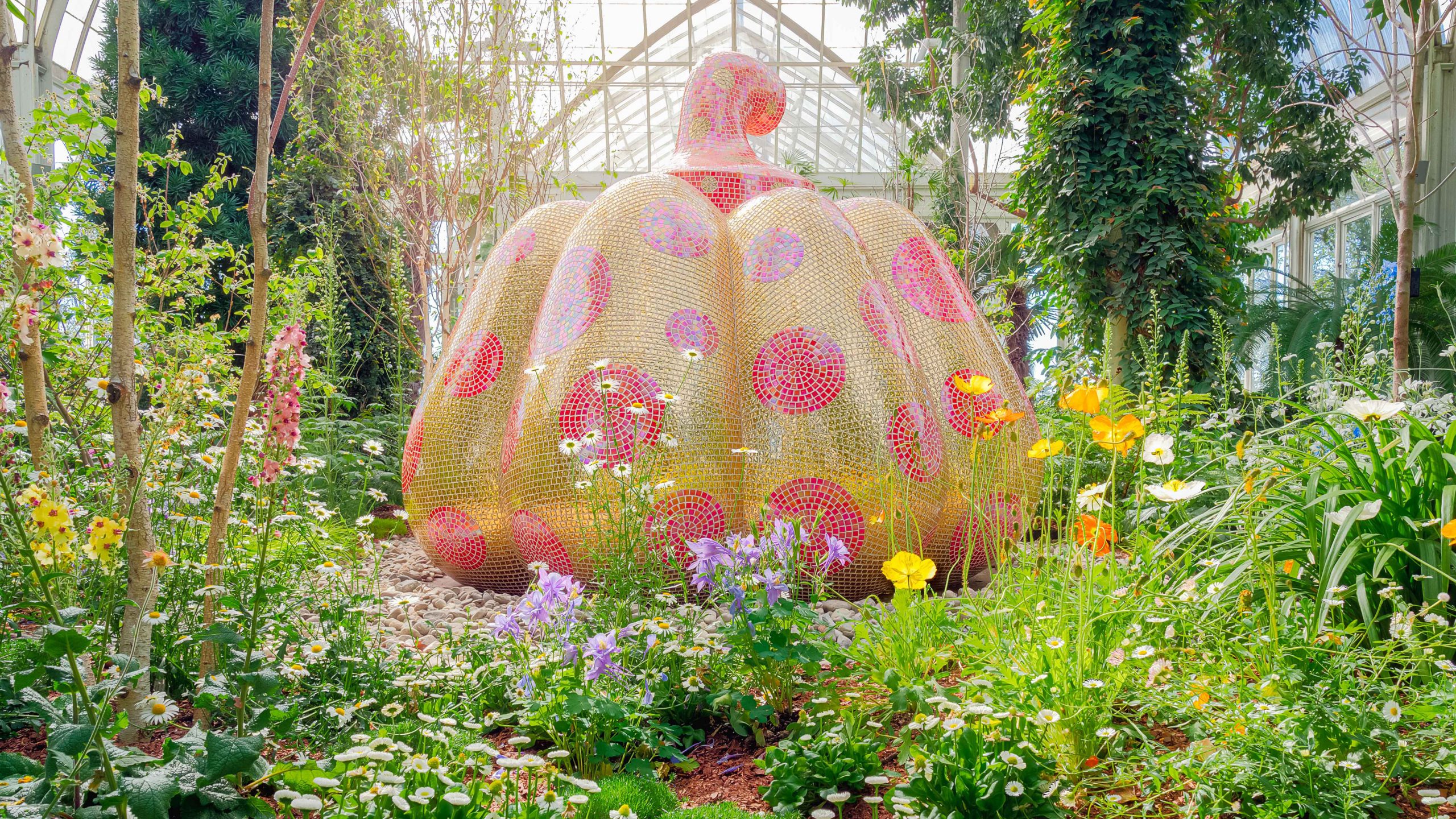 A large pumpkin sculpture covered in yellow mosaic tiles with red mosaic polka dots. Its stem is covered in red mosaic tiles with small yellow mosaic polka dots. The sculpture is nestled into a bed of low-lying flowers and plants, with small trees and vines rising up just behind it.
