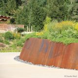 A concrete path curves around a raised, tilted piece of rusted steel, within which sits a planting of diverse flowering shrubs; a forest is visible in the background, along with the eaves of a house