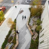 A view from above of an urban greenspace, composed of a slanting cobbled path lined with granite stones, trees, shrubs, grasses, and integral benches