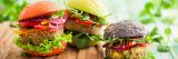 Plant-based burgers in a variety of shapes and colors sit on a cutting board, plants visible in the background