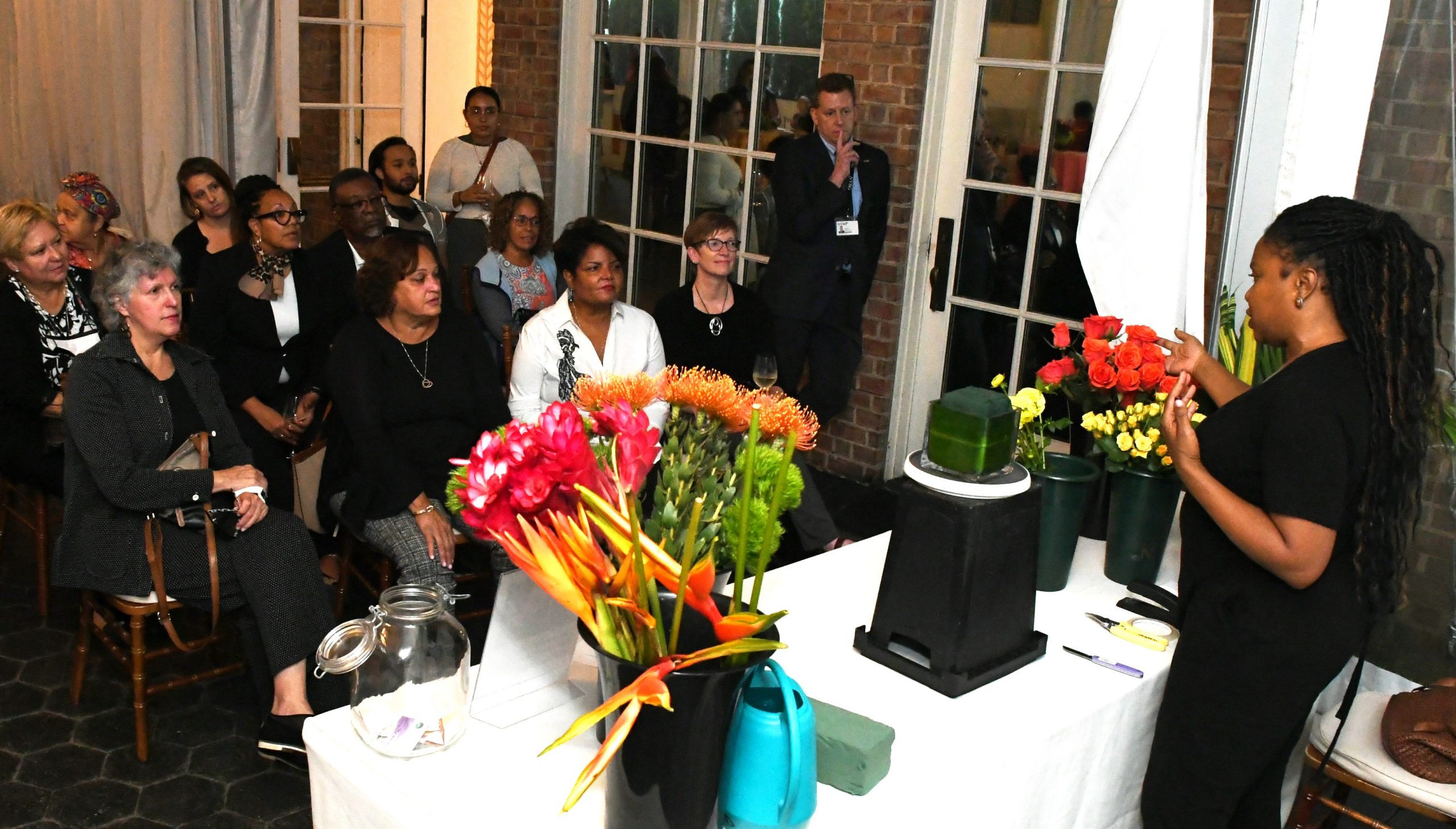 Guests sit for a floral design demonstration by a woman in black, standing behind a table covered in vases and flowers