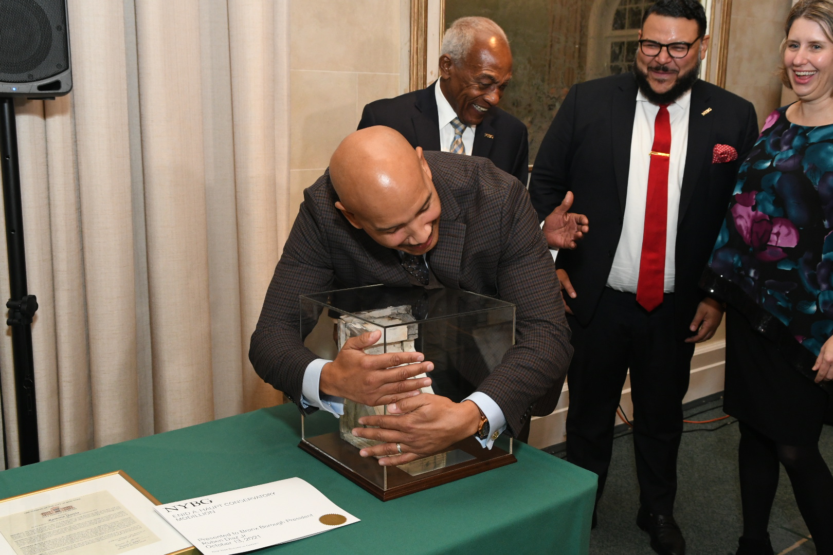 A man in a suit, backed by other attendees of a formal event, jokingly hugs an award he received