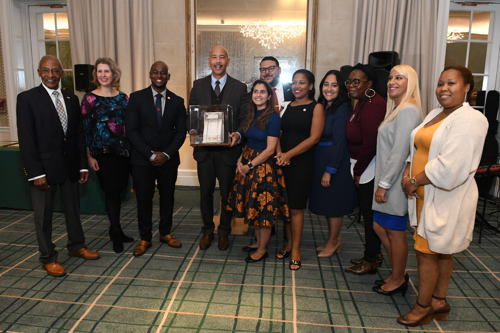 An award recipient stands at the center of a group of people dressed for an evening event, displaying his award for the photographer