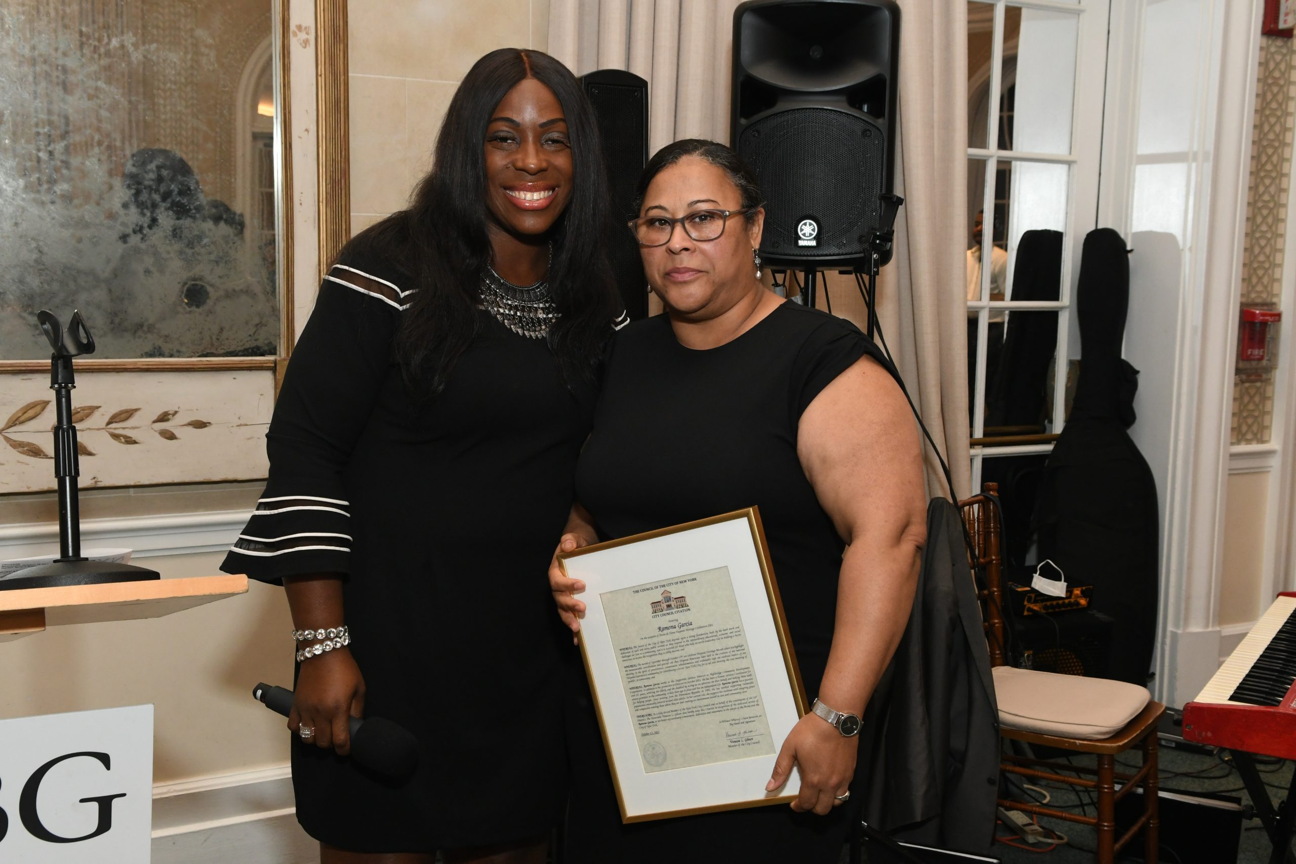 Two women in black outfits pose with a framed certificate