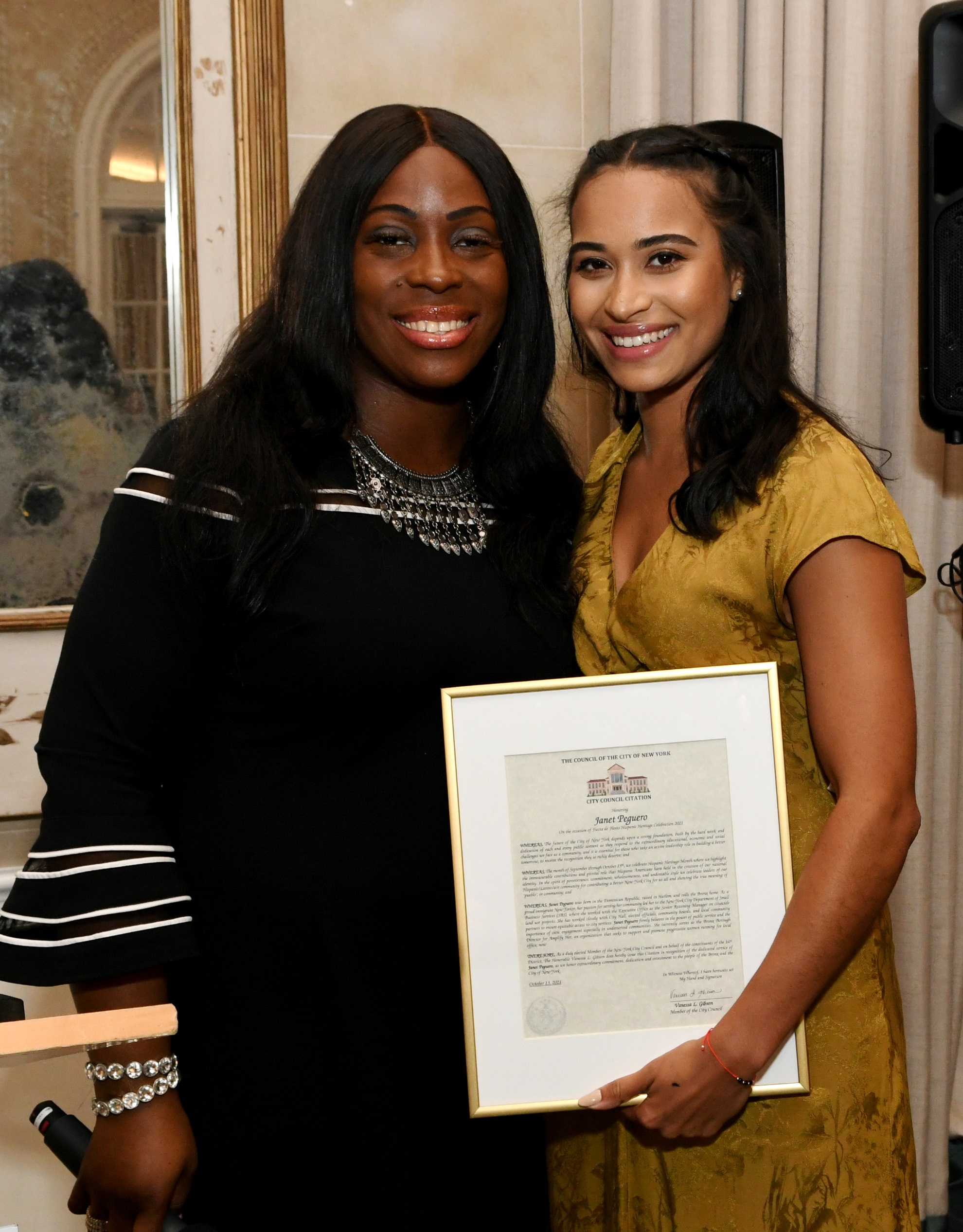 Two women in black outfits pose with a framed certificate