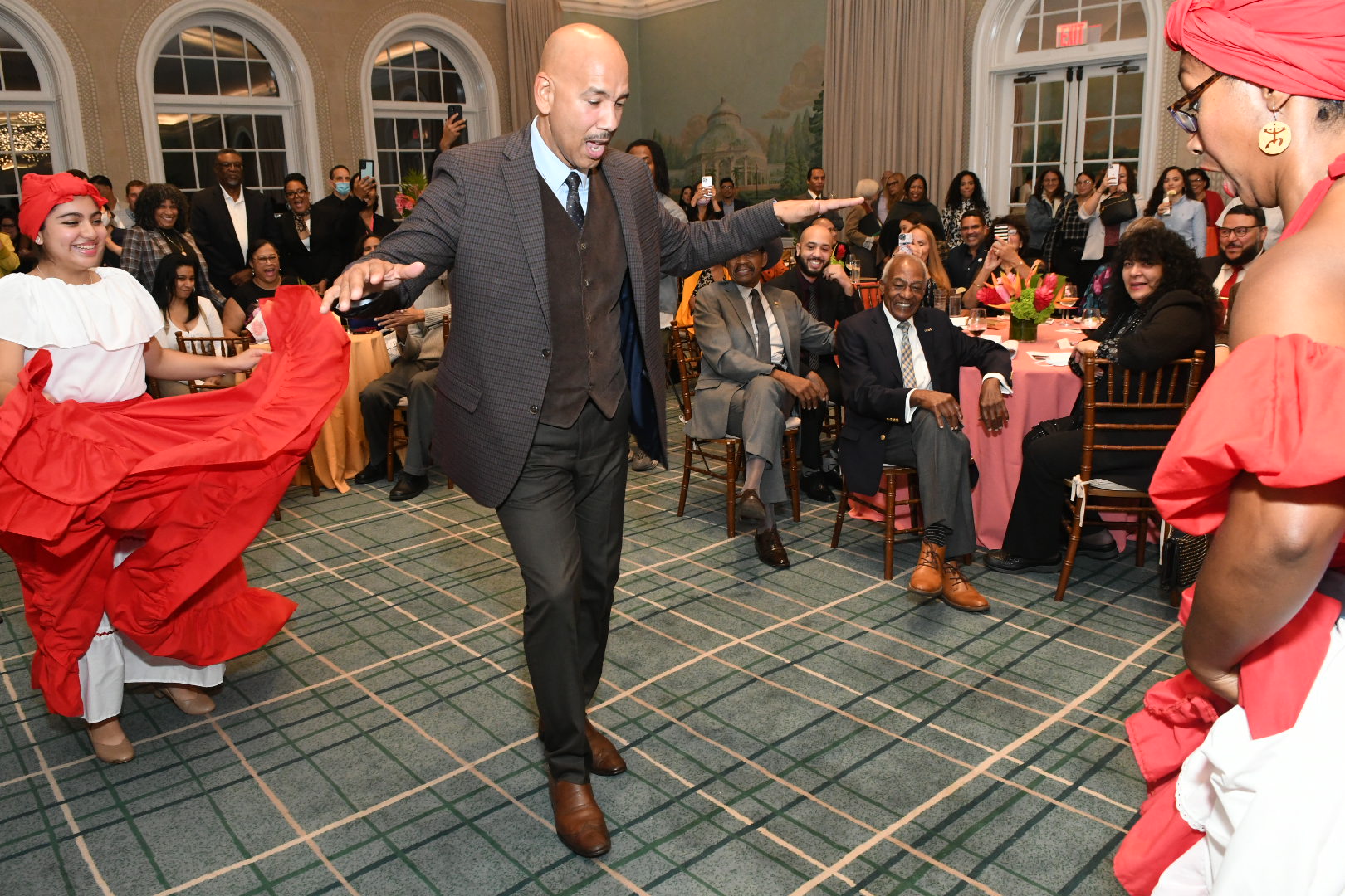 A man in a suit, surrounded by women in flowing red dresses, dances to live music