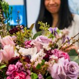 A woman with long, dark hair works on a rose-themed floral arrangement of pinks and purples