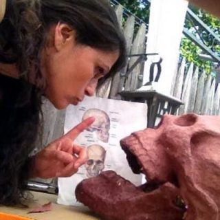 A dark-haired woman leans over a clay skull, wagging her finger at it