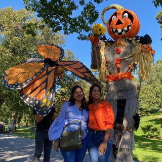 Two women in brightly colored shirts pose in front of tall, human-controlled puppets, one a monarch butterfly and the other a scarecrow