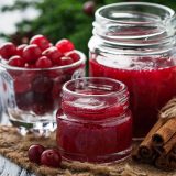 Three clear glass jars sit arranged on a table, full of cranberries and cranberry jam and surrounded by ingredients