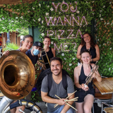 A brass band of five people poses with their instruments in front of a sign that says "You wanna pizza me?"