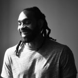 A person in a T-shirt with long dreadlocks poses for a black and white photo in the sunshine