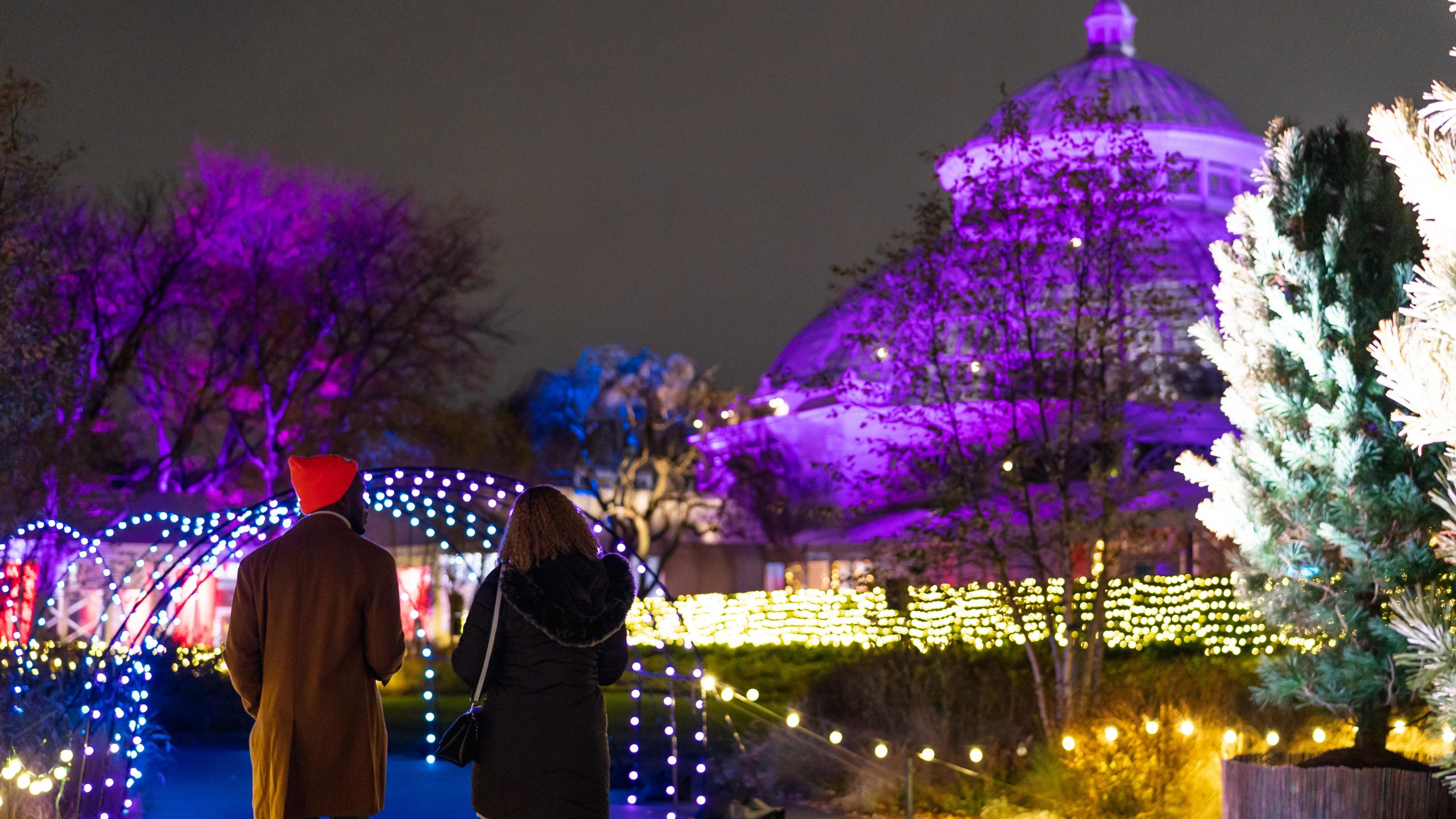 A couple walking down a pathway with a row of lights in front of the illuminated Conservatory