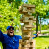 In focus is a JENGA tower out of focus is a person wearing all blue opening arms happily