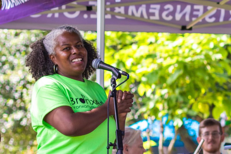 A woman in a bright green shirt, lips upturned, speaks into a microphone at a sunny event