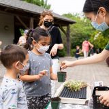 Two small children engage with a young woman in a mask as they learn how to plant a seedling in a pot of soil