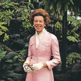 A woman with brown hair and a pink coat poses for a photo among tropical foliage