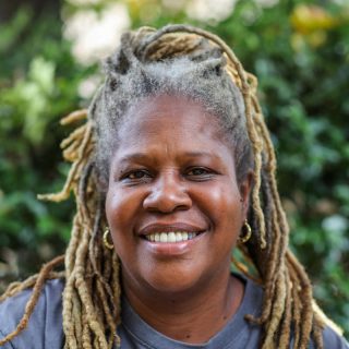 A woman with dreadlocks and a gray shirt poses for a photo in front of a sunny garden scene