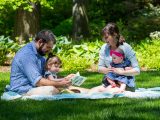 family of four sitting on a picnic blanket, one of the parents is reading to the family
