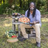 A man with long, dark braided hair, wearing a purple shirt and khaki pants, displays a plate of shrimp and vegetables in a natural setting