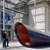 Cut at a bias, a large, black pipe lined on the interior with slabs of red-stained wood sits in the sunshine while two people observe