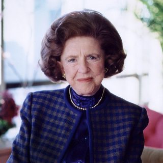 A woman with brown hair poses for a photo wearing a blue and black coat and blouse