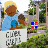wooden sign of a woman and child holding a globe in the forefront and international garden beds in the background