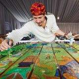 Artist wearing a bright red and orange had with a white shirt, leaning over colorful a table painting people and green leaves and birds. Holding a paint brush in one hand looking at the table, and a paint pallete in the other hand with a cup of water and another paint brush close to the table.