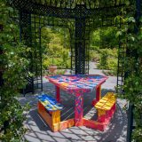 Colorful pink, blue and yellow painted wooden picnic table placed unter a dark green gazebo structure with green leaves winding all around it.