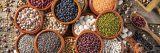 Assorted legumes in round clay containers on rustic wooden background and wood serving scoop with mixed legumes including lentils, chickpeas, soybeans and beans