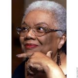 Headshot of Lucille Clifton, with short cropped grey hair, wire rimmed glasses, red lip, and face is resting on a hand, wearing beaded dangling earrings.