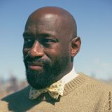 Headshot of Dante Michaeux, wearing a yellow bowtie, white collared shirt, and tan sweater, black beard and mustache, with a bald head, against a blured blue sky line.