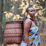 A woman in a blue wrap dress and large shell earrings looks at the camera as she walks with a basket of foraged plants worn on her back