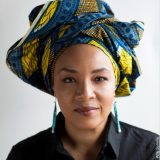 Headshot of LaTasha N. Nevada Diggs, wearing a black collared shirt, long turquise earrings, and a blue and yellow colorful headwrap.