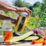 A pair of hands works to open a jar of freshly packed pickles over a table of sliced green cucumbers, orange and red peppers, pink and yellow flowers, and a background of green foliage on a sunny day.