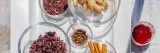 Top down view of glass bowls with dried red rose petals, cinnamon sticks, ginger on a white plate, next to a paper cup with a red liquid.