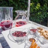 Variety of glass bowls with dried red rose petals, cinnamon sticks, ginger on a white plate, next to a glass pitcher with a red liquid.