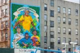 A colorful mural in blues, yellows, and greens covers the front of a gray building, depicting a woman with long gray dreadlocks, arms opened and lips upturned. Three additional people in colorful clothing are arrayed beneath her, working with fruits and vegetables. It says "Garden of Happiness" on a painted banner across the top.