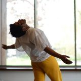 A man in a white shirt and yellow pants performs an interpretive dance in a sunny room