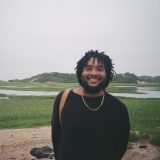 A man in a black shirt with upturned lips poses for a picture before a coastal wetland environment