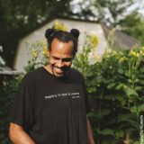 Headshot of Ross Gay standing next to tall green plants with yellow flowers and a barn in the background. Wearing a black shirt.