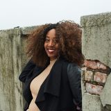 A woman in a black jacket and tan sweater, with lips upturned, poses for a photo among old architecture