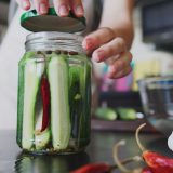 Clear jar with slivers of cut open green cucumbers in liquid with red chilis representing pickles.